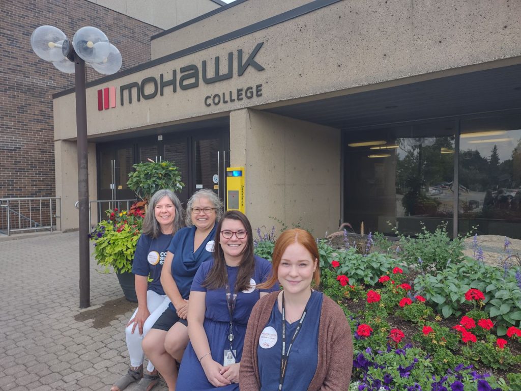 Pictured right to left is Sara, Caitlin, Melissa and Tracey-Ann. They are seated in front of flowers at the main doors to the Mohawk College C wing