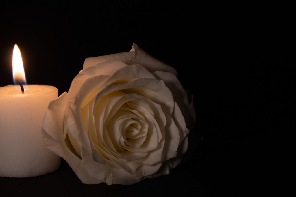 A solitary white rose sits on a black background with a lit white candle